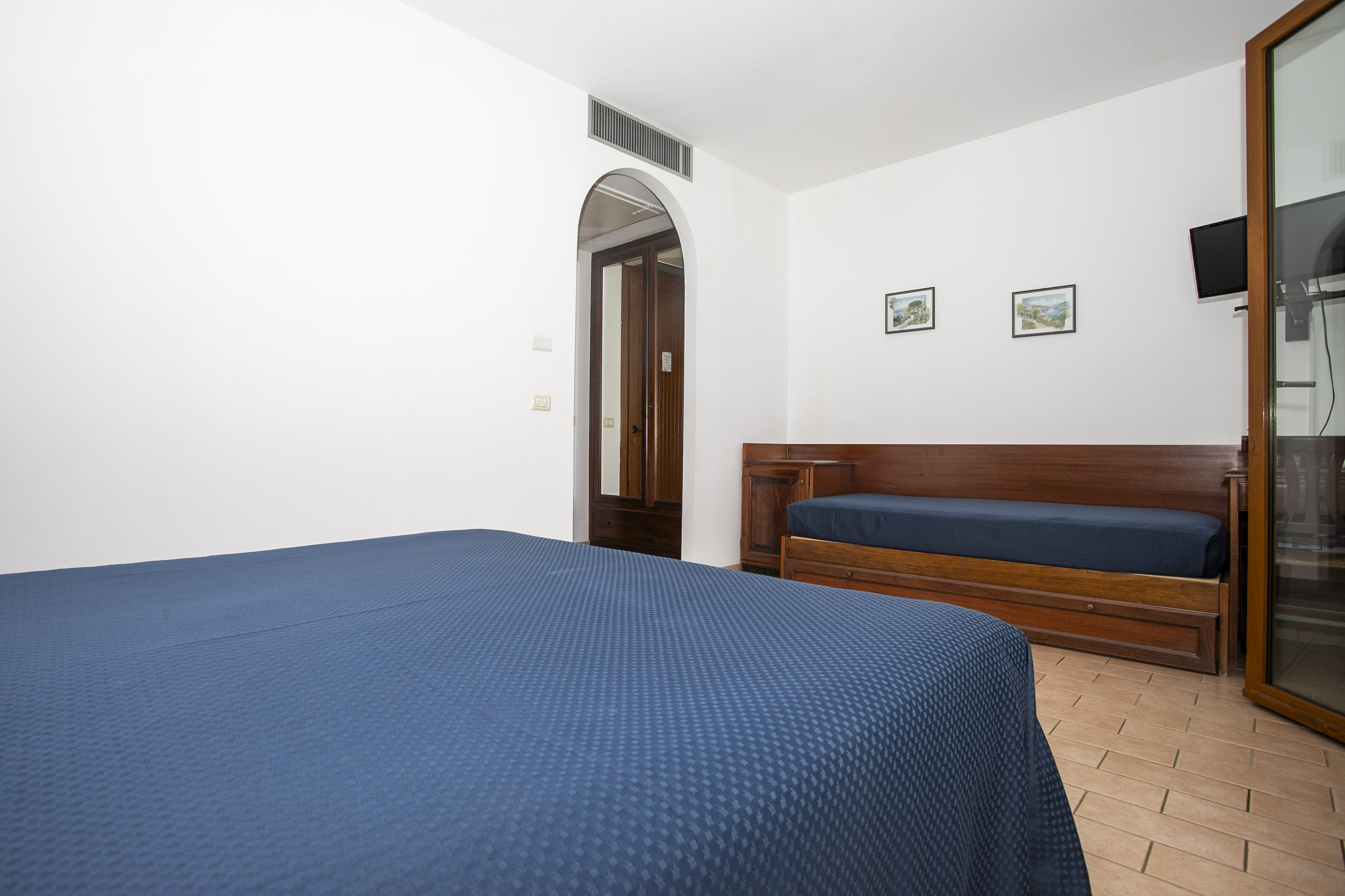 A hotel bedroom with a blue bed, wooden furniture, terracotta tiles, a TV, and a doorway to another room or hallway. The r...